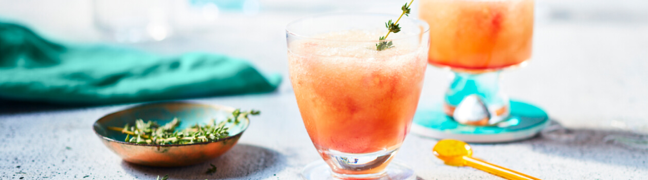 Glasses filled with a nectarine-hued Slushy Shirley cocktail garnished with a sprig of thyme sitting on a gray countertop.